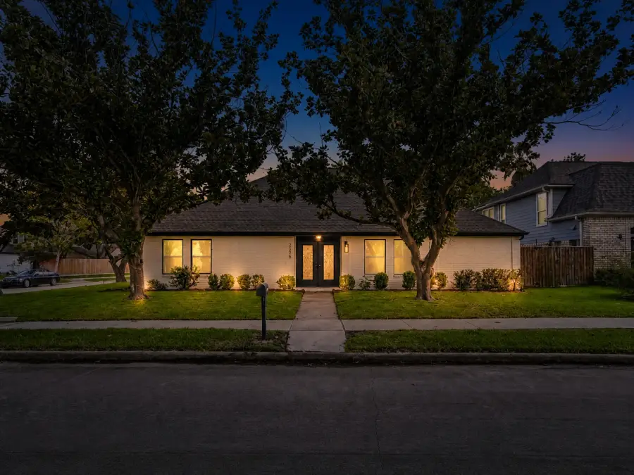 After AI day to dusk conversion, the same white-brick home at dusk with a deep blue sky, glowing windows, lit entry and landscape lighting, with the roofline, trees and lawn unchanged